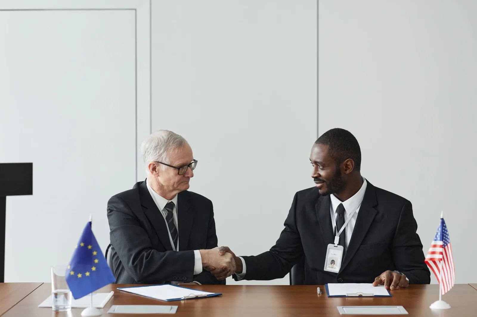 Corporate handshake between diverse businessmen representing EU and US flags, symbolizing partnership and collaboration. Stock photo illustrating Craftroot Retail's regulatory approval story.