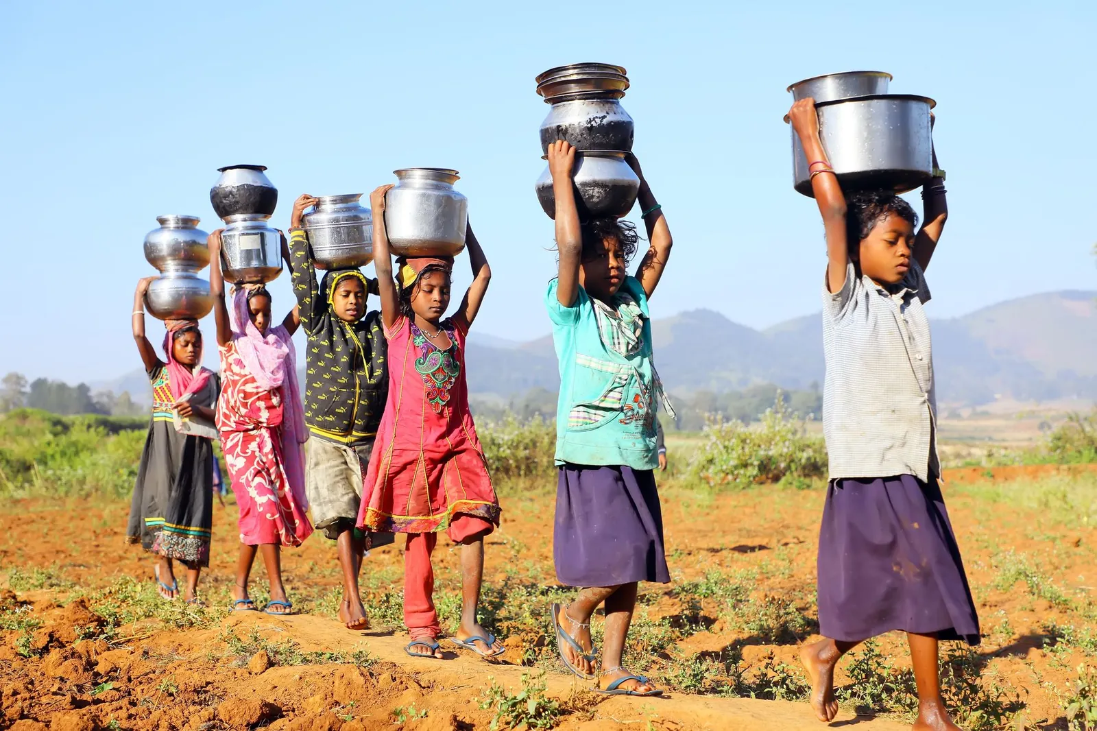 A group of children carry stainless steel pots on their heads in a rural setting, showcasing daily life. Stock photo illustrating Satin Creditcare Network's fund raise story.