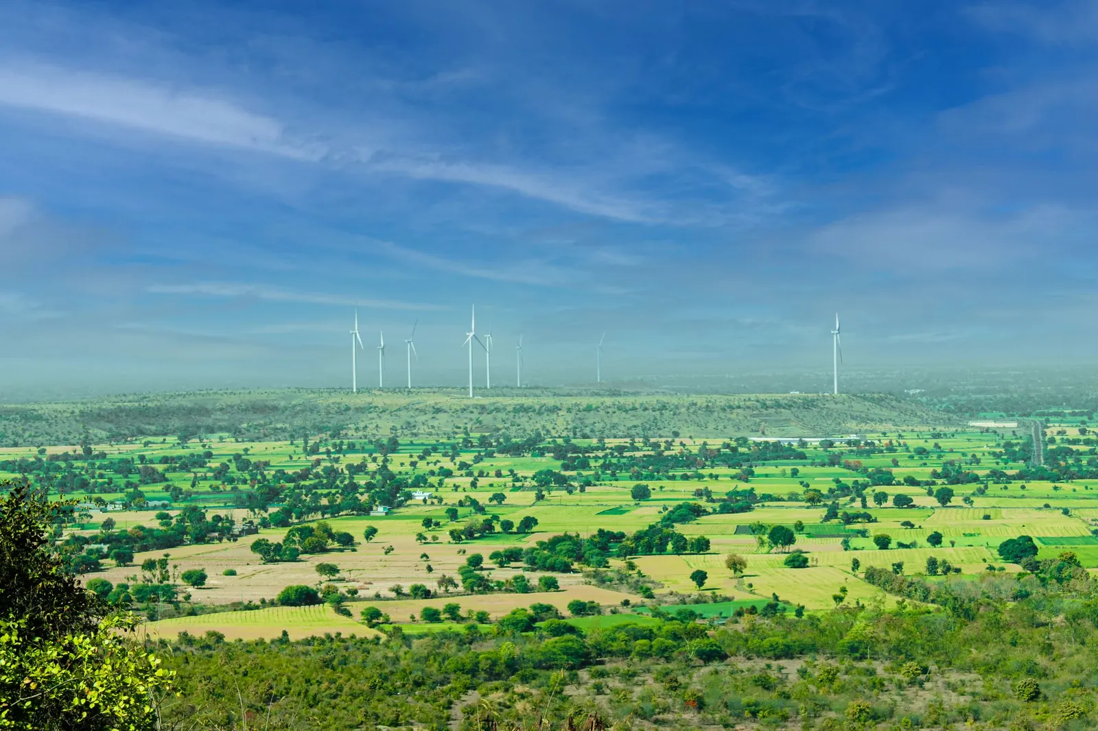 A stunning view of wind turbines towering over vibrant green fields under a clear blue sky in Rajapur, India. Stock photo illustrating Enviro Infra Engineers' new order story.