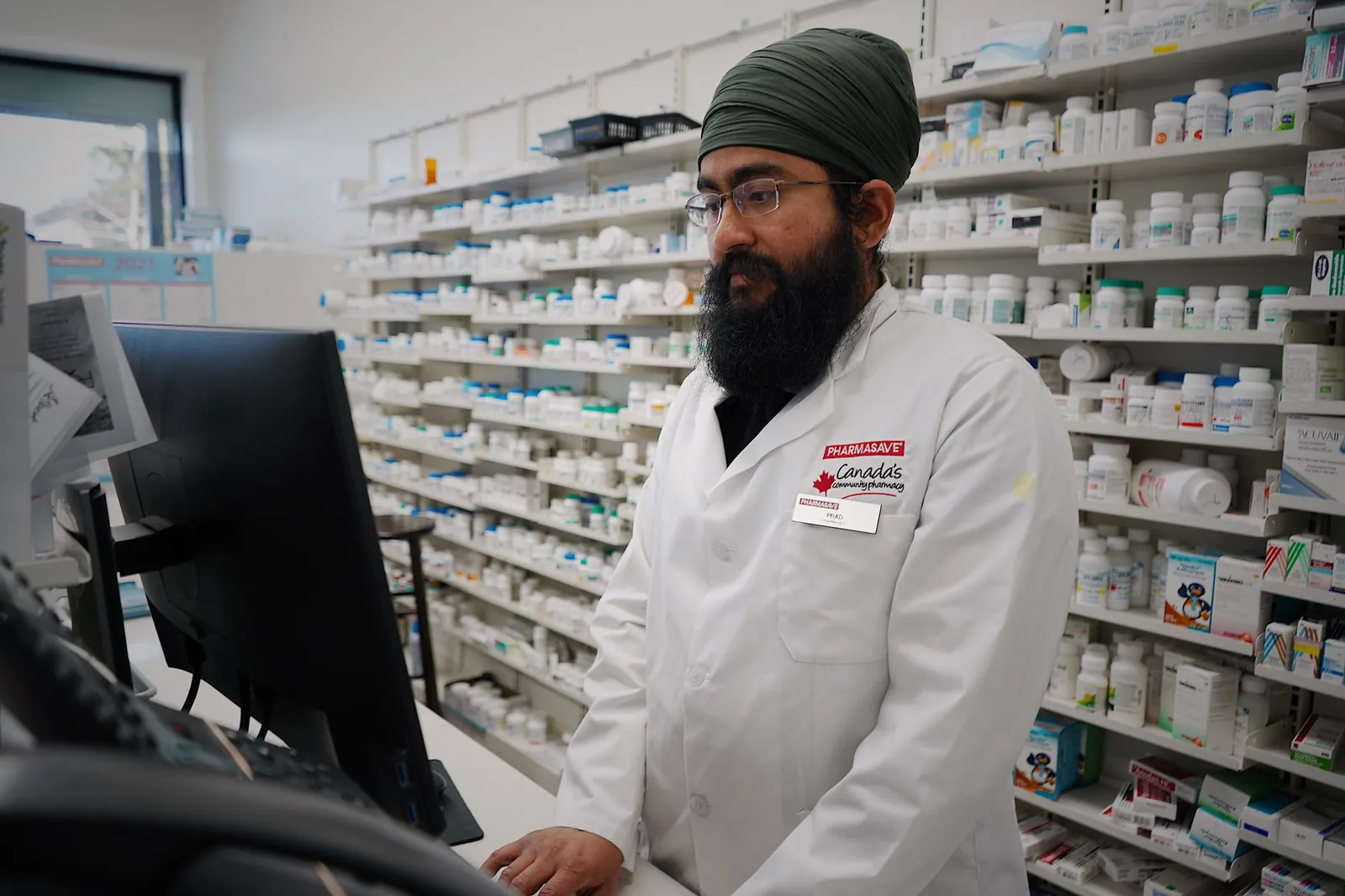 Pharmacist wearing a turban working at a computer in a pharmacy store with shelves filled with medicine. Stock photo illustrating Zota Health Care's expansion story.