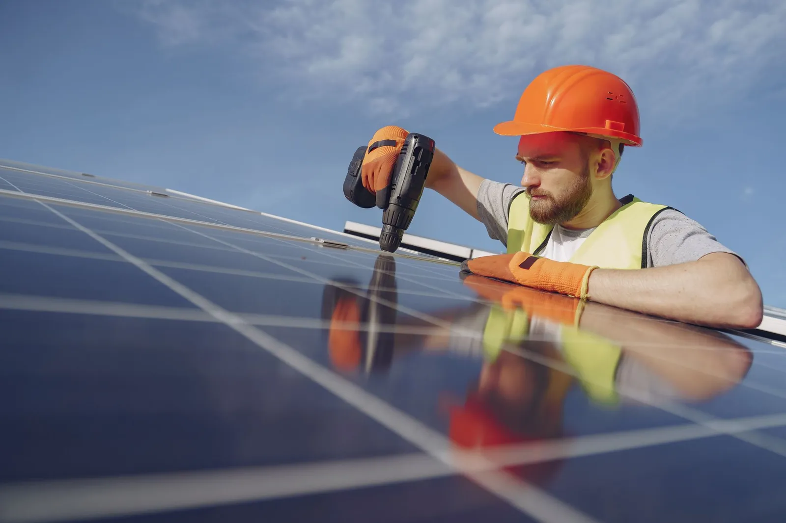 Low angle of serious bearded male electrician in hardhat and protective gloves installing solar photovoltaic panel system using drill. Stock photo illustrating Saatvik Green Energy's new order story.
