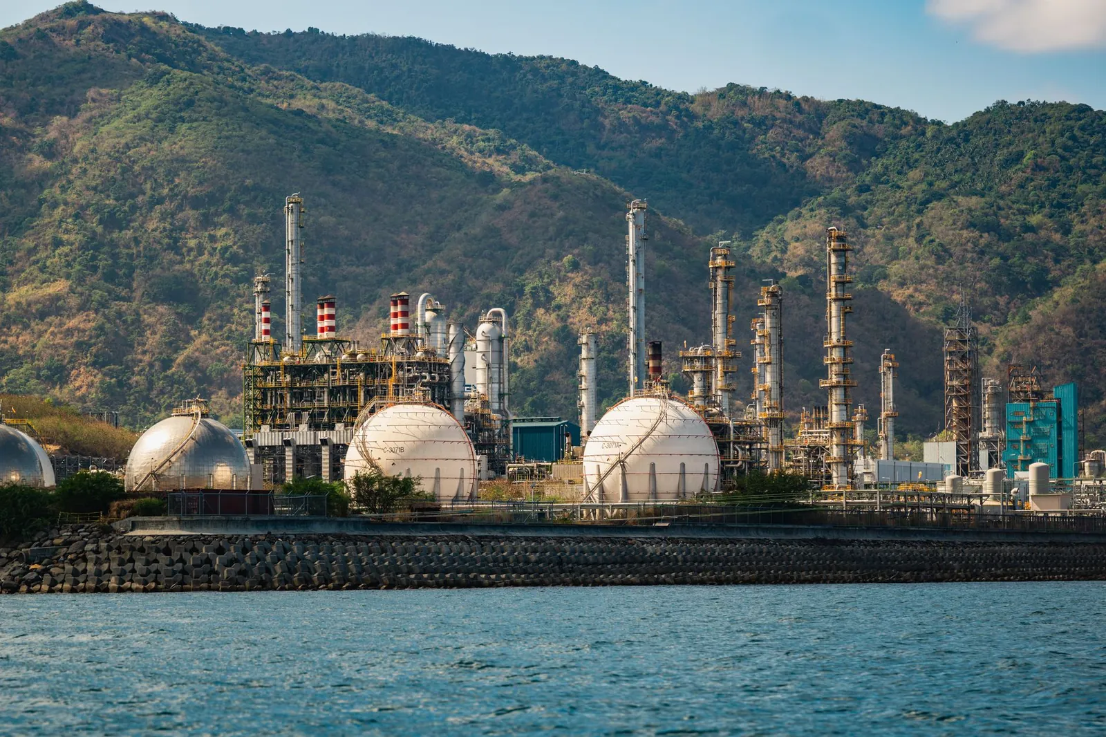 Industrial refinery with spherical tanks by the coastline against a backdrop of mountains. Stock photo illustrating Hindustan Petroleum Corporation's production started story.
