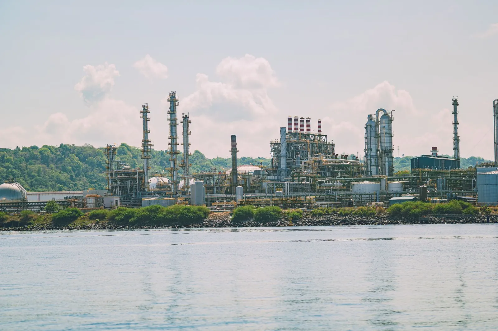 A large oil refinery situated by the water with bright green foliage in the background; clear blue skies and clouds overhead. Stock photo illustrating GP Petroleums' fund raise story.