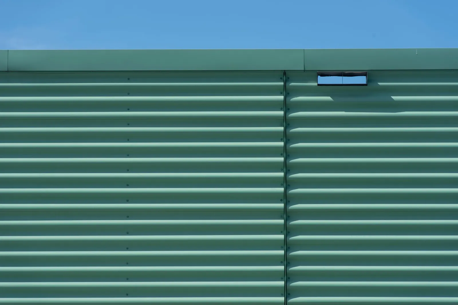 Minimalistic view of a green metal wall with horizontal panels against a clear blue sky, captured outdoors. Stock photo illustrating HLE Glascoat's new order story.
