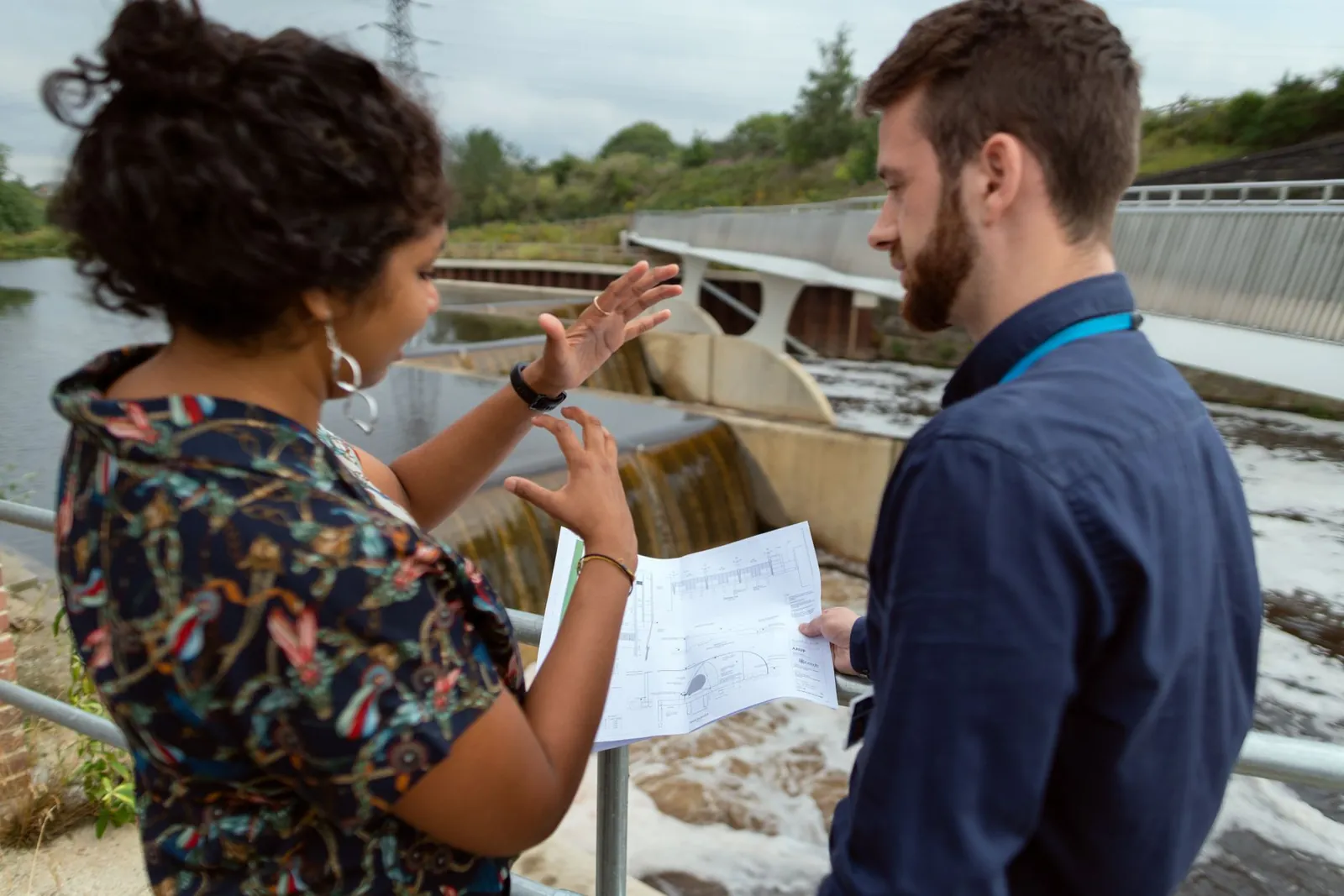 Two engineers discussing plans by a water project site. Collaborative teamwork on hydropower. Stock photo illustrating Univastu India's earning call story.