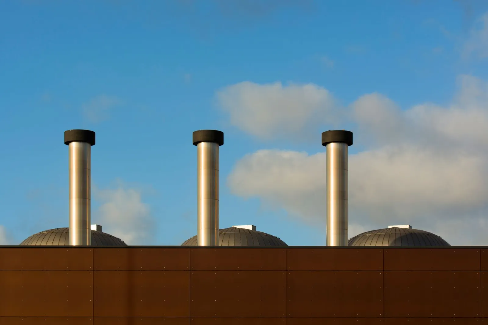 Three industrial chimneys reach into a serene blue sky dotted with clouds. Stock photo illustrating GHV Infra Projects' new order story.