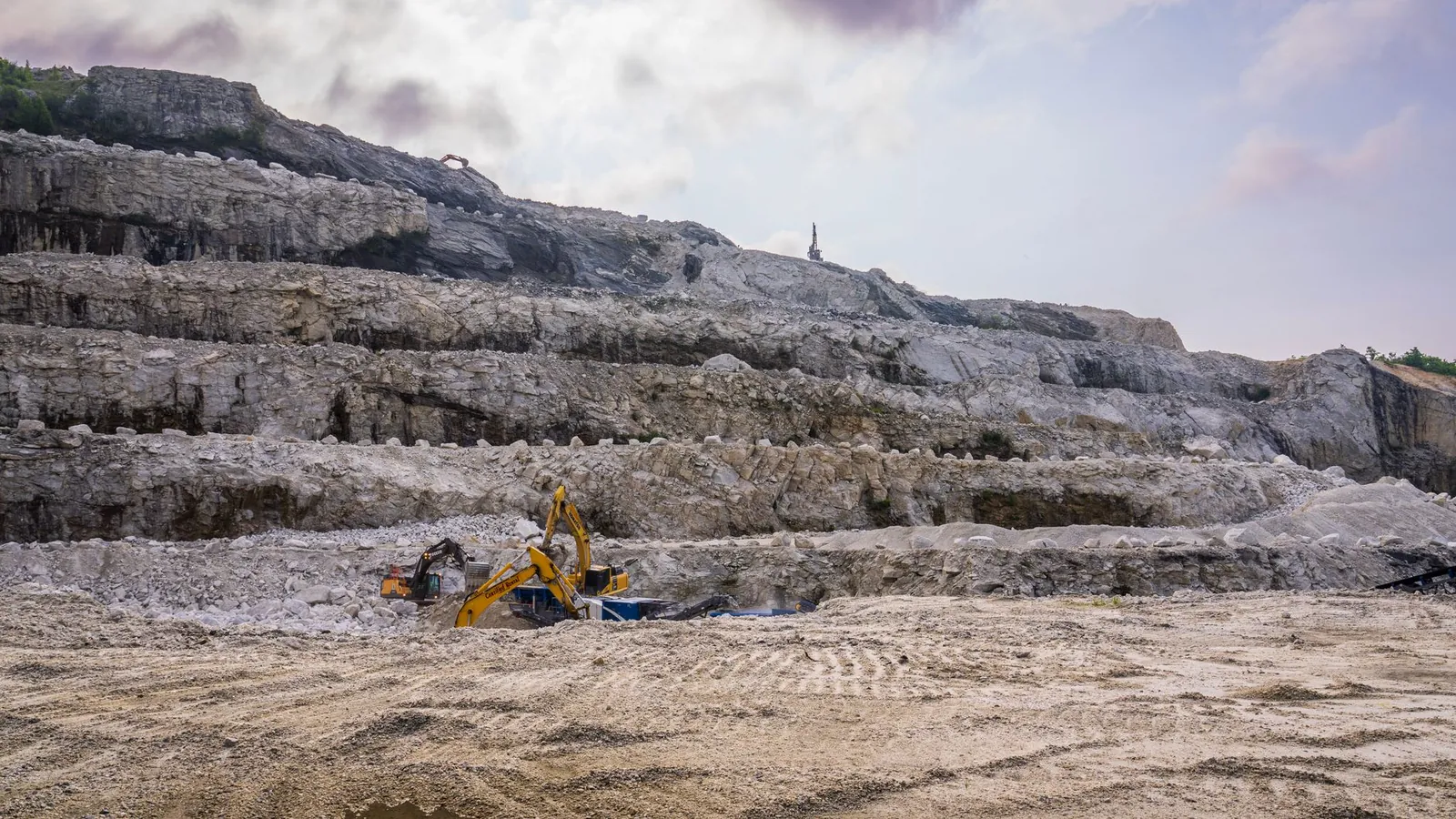 A quarry with excavators working on rocky terrain under a cloudy sky. Stock photo illustrating Shiva Cement's expansion story.