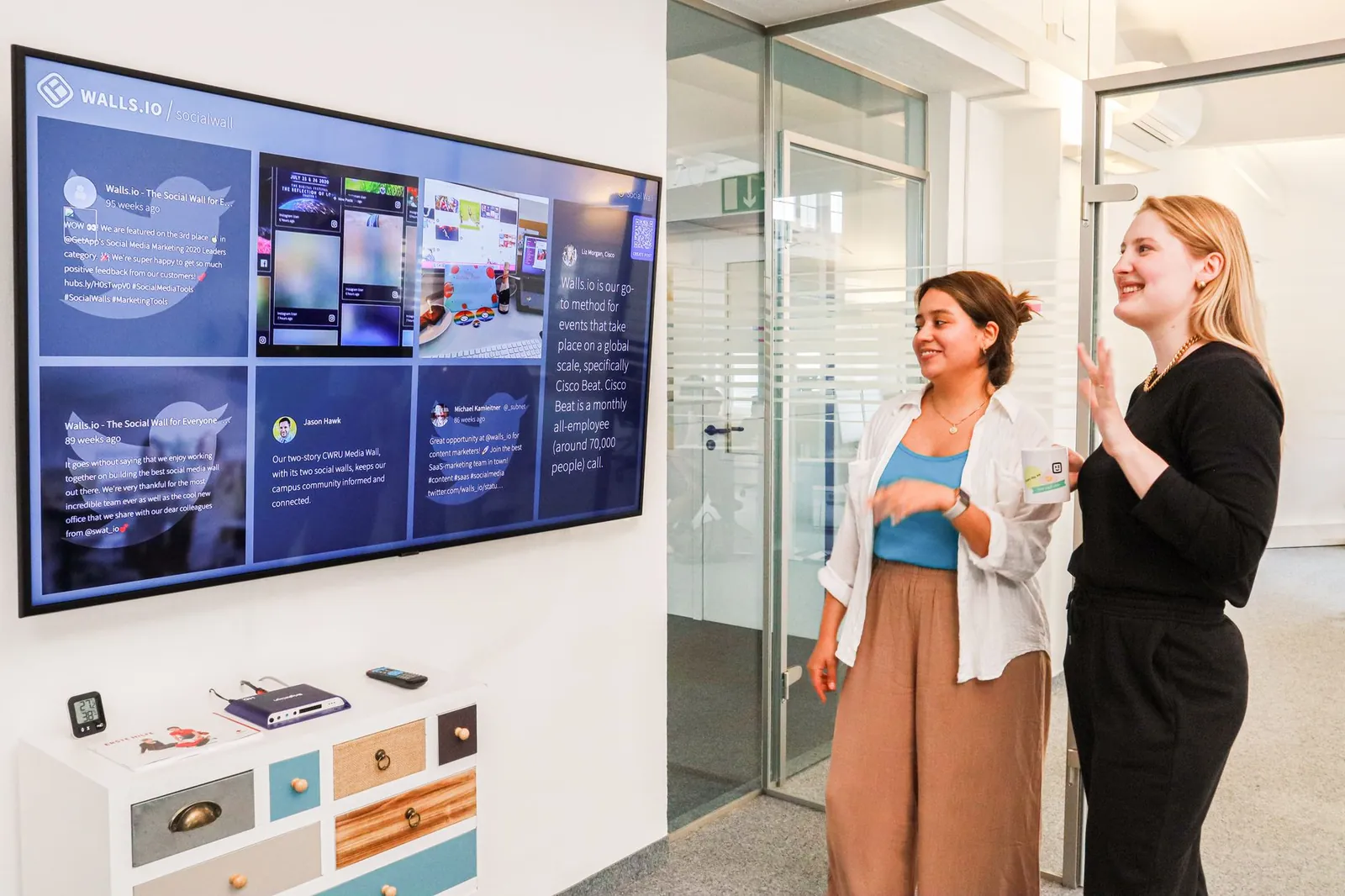 Two women in an office environment interact with a large digital display showcasing social media content. Stock photo illustrating Quint Digital's promoter buying story.