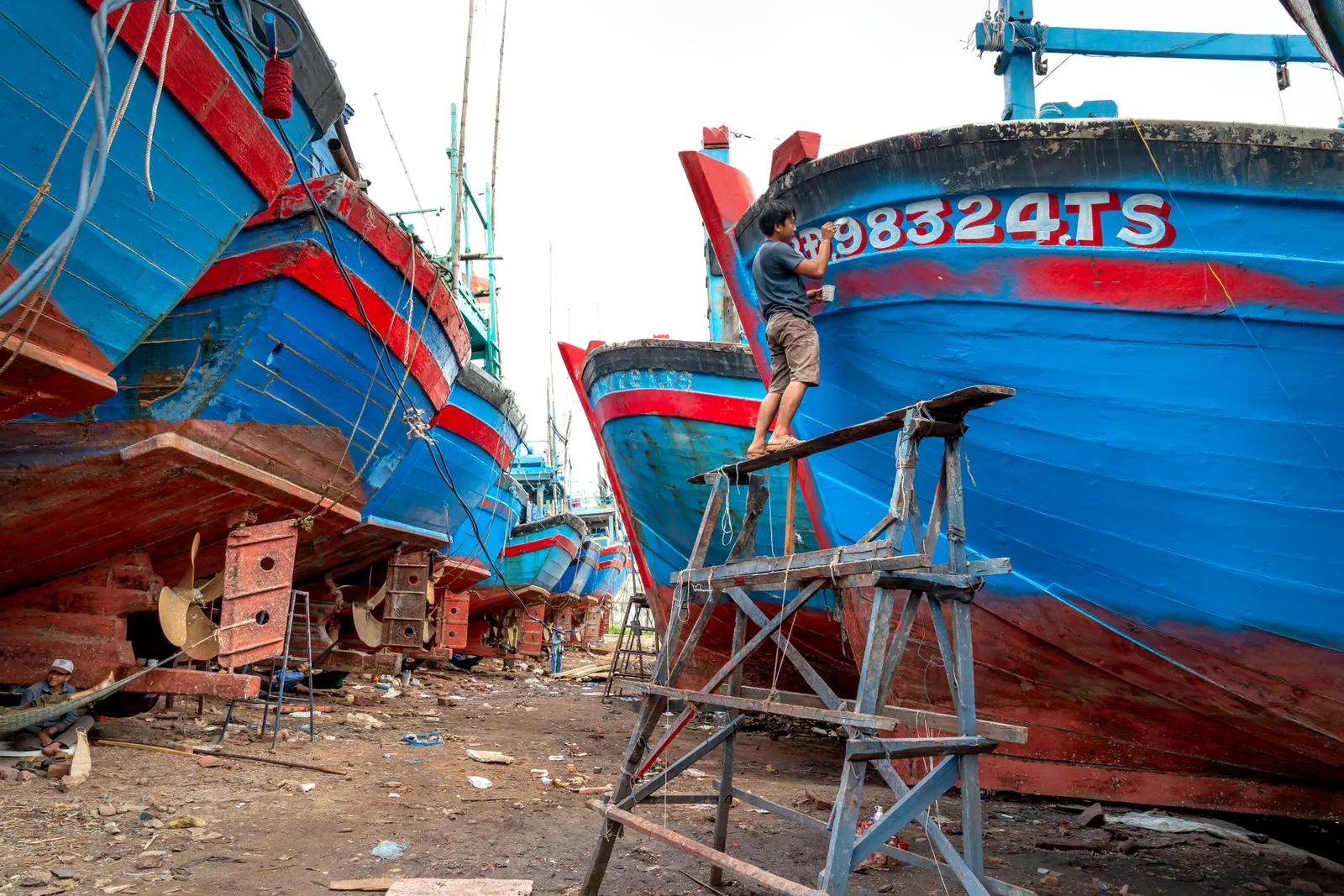 A man stands on scaffolding, painting a large wooden ship in a dry dock area. Stock photo illustrating Cochin Shipyard's expansion story.