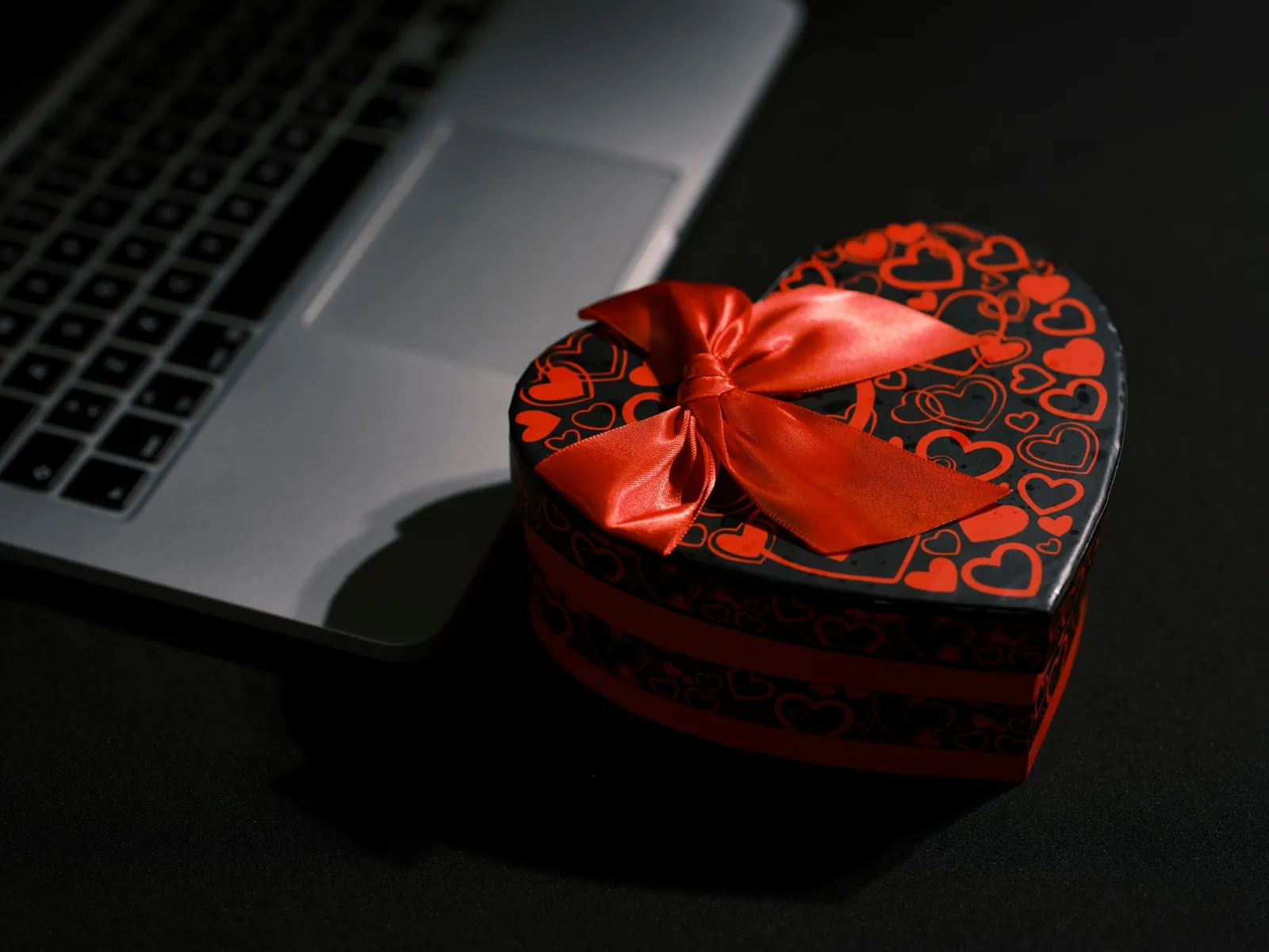 Heart shaped black and red gift box with ribbon next to a laptop on a dark table. Stock photo illustrating 360 ONE WAM's regulatory approval story.