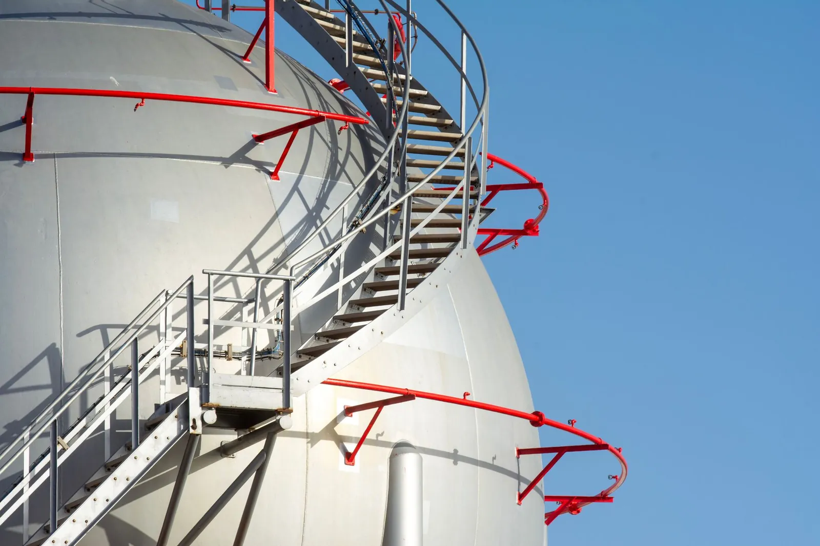 Close-up of a large industrial storage tank with spiral steel stairs against a clear blue sky. Stock photo illustrating Desco Infratech's partnership story.