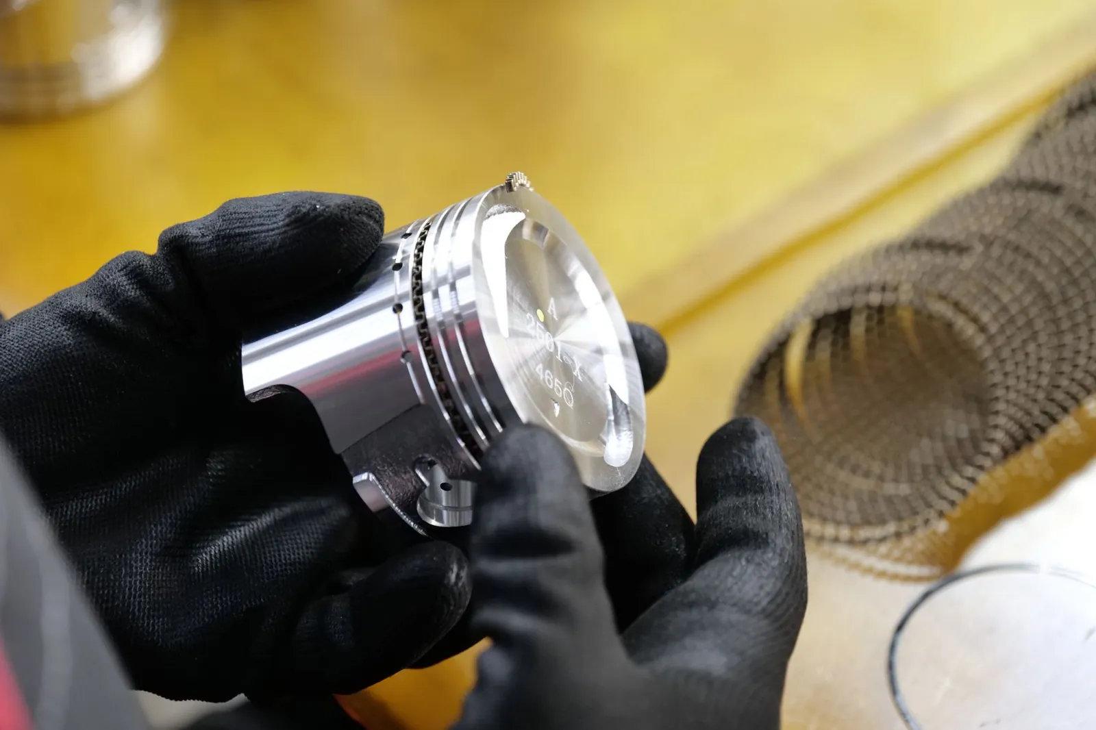 Close-up of black-gloved hands examining a metal engine piston in a workshop. Stock photo illustrating IP Rings' expansion story.