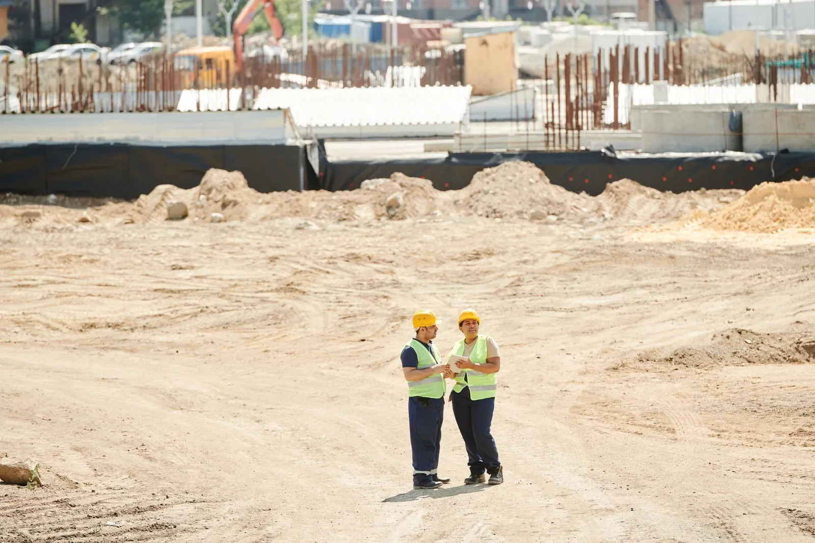 Two workers discussing plans on a sandy construction site, wearing safety gear. Stock photo illustrating Dilip Buildcon's new order story.